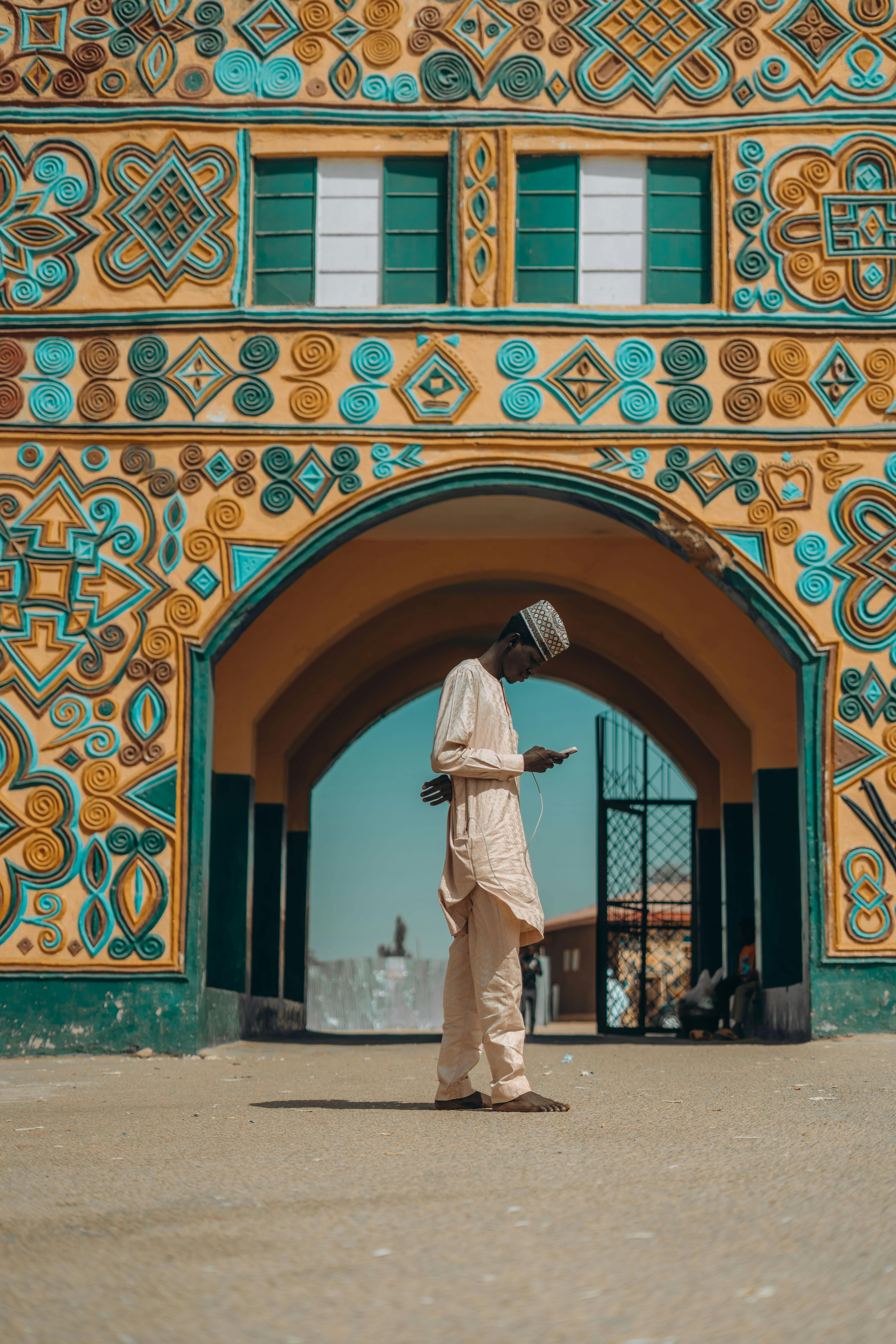 Man in traditional attire standing barefoot, looking at phone under an arch with colorful patterns.