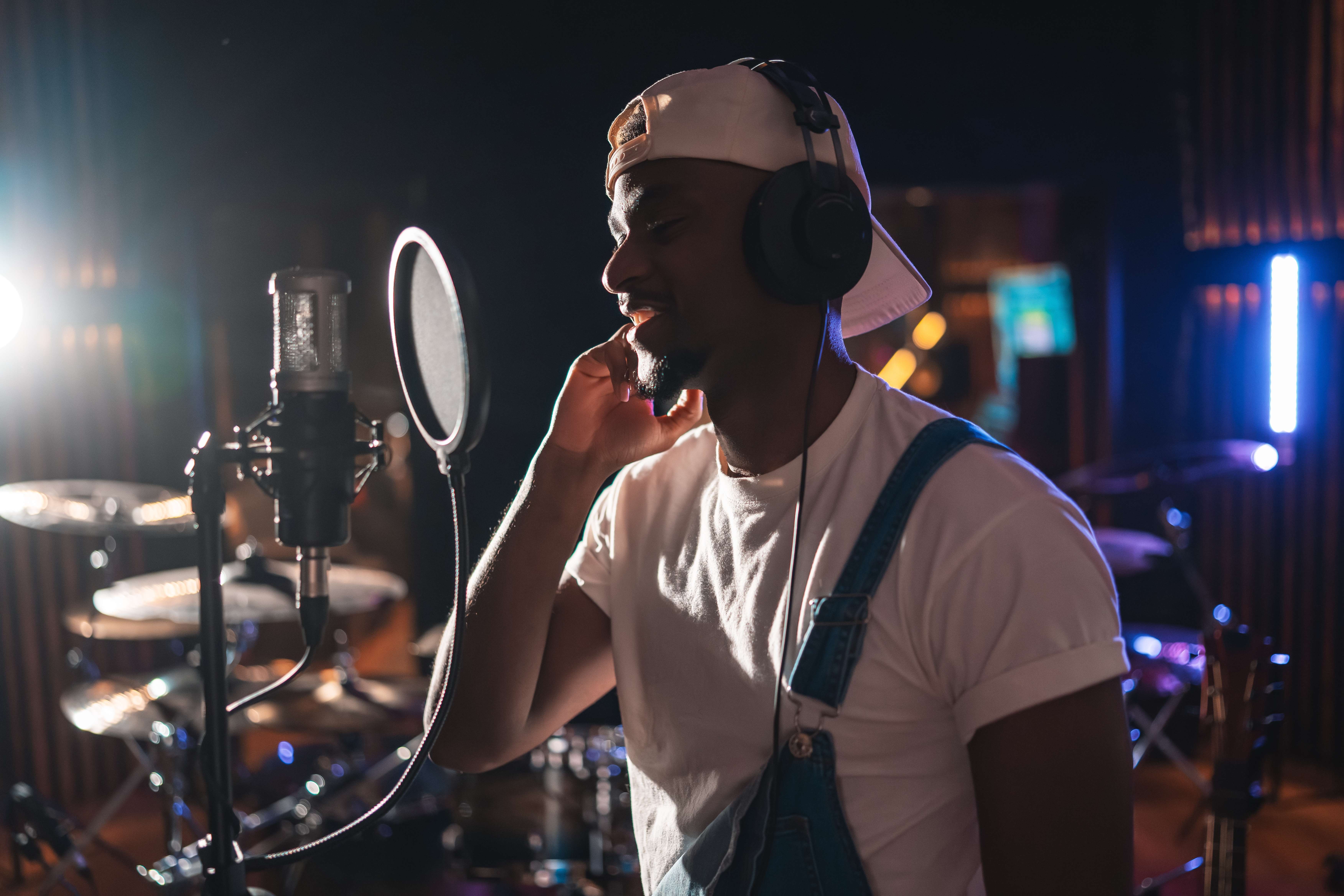 Man wearing headphones and a backward cap recording vocals in a music studio near a microphone.